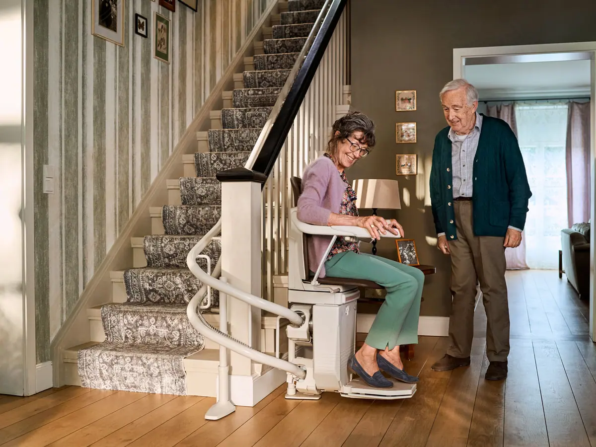 Smiling older woman seated on a GlideWell straight stairlift while a family member stands beside her on the staircase